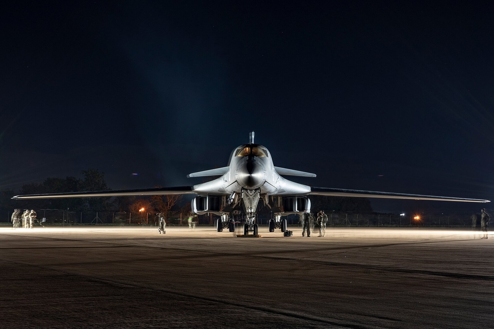 夜間燈光下，一架 B-1B 轟炸機正面停在英國空軍基地停機坪上，可變後掠翼展開，地勤人員在機旁作業