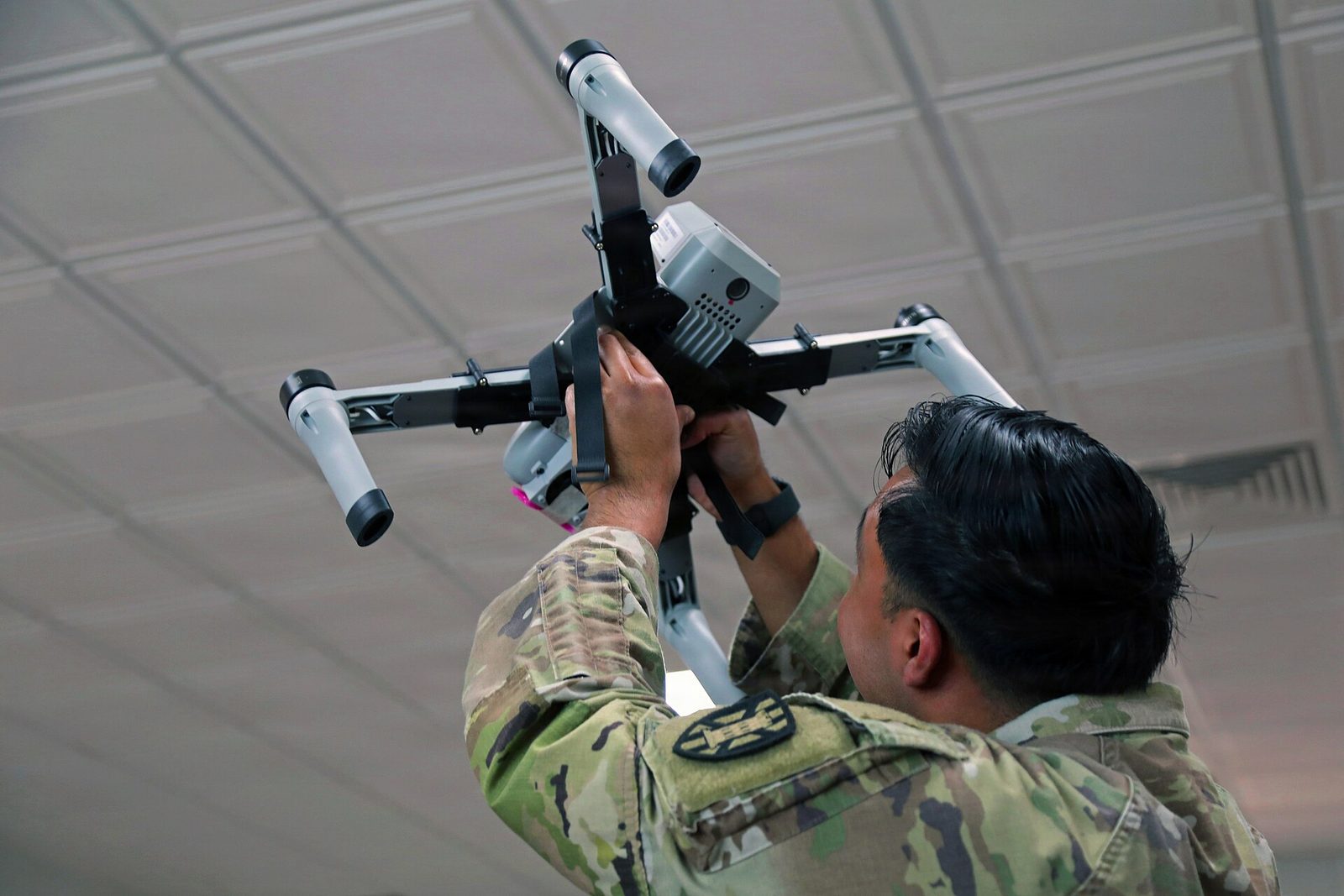 一名身著迷彩服的美軍士兵雙手高舉一具四旋翼反無人機系統，仔細檢視機體與酬載