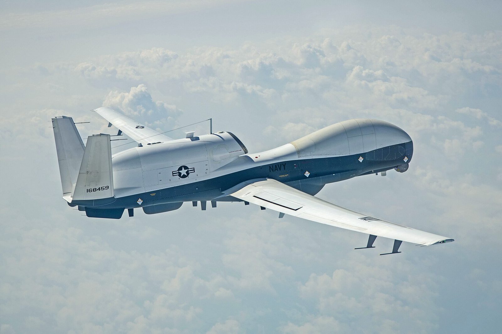 一架白色機身、藍色腹部的 MQ-4C Triton 高空長航時無人機在雲層上方飛行